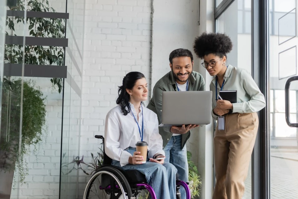 Group of 3 people, one woman disabled in wheelchair, all looking at laptop together